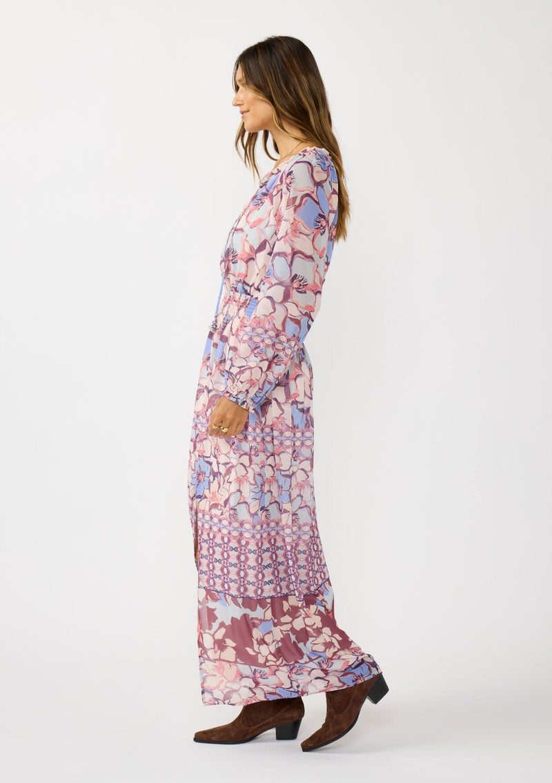 [Color: Natural/Dusty Blue] A side facing image of a brunette model wearing a blue and pink floral maxi dress with long sleeves, a split v-neckline with ties, a smocked waist, and a side slit. 
