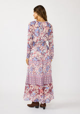[Color: Natural/Dusty Blue] A back facing image of a brunette model wearing a blue and pink floral maxi dress with long sleeves, a split v-neckline with ties, a smocked waist, and a side slit. 