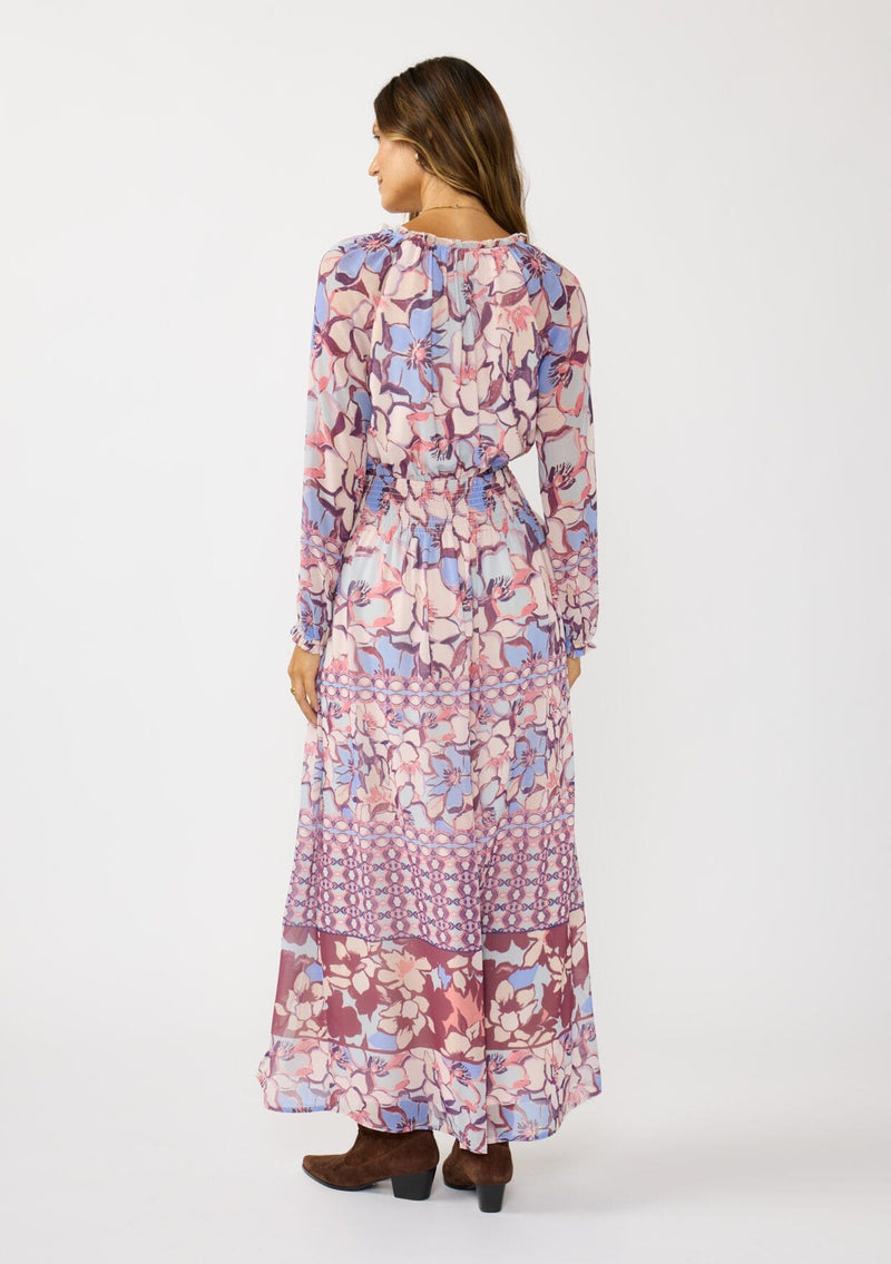 [Color: Natural/Dusty Blue] A back facing image of a brunette model wearing a blue and pink floral maxi dress with long sleeves, a split v-neckline with ties, a smocked waist, and a side slit. 