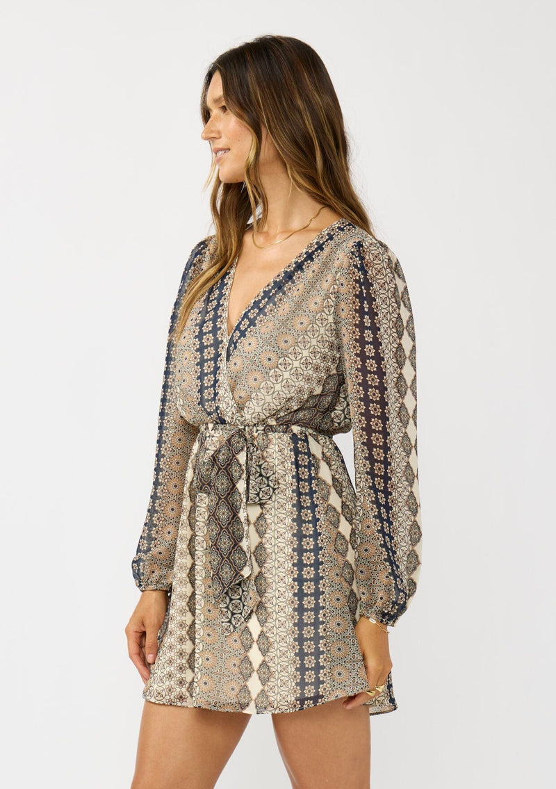 [Color: Natural/Navy] A side facing image of a brunette model wearing a striped floral pattern mini dress with long sleeves, a surplice v-neckline, and an elastic waist with tie detail. A casual fall boho dress for brunch and special occasions. 