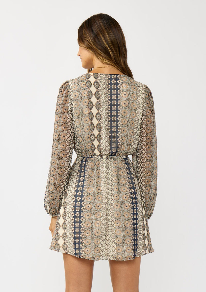 [Color: Natural/Navy] A back facing image of a brunette model wearing a striped floral pattern mini dress with long sleeves, a surplice v-neckline, and an elastic waist with tie detail. A casual fall boho dress for brunch and special occasions. 