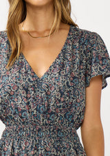 [Color: Navy/Rose] A detail image of a brunette model wearing a navy floral border print midi dress with a classic v-neckline, short sleeves, a smocked waist, and side pockets. A fall bohemian dress perfect for any occasion. 