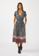 [Color: Navy/Rose] A front facing image of a brunette model wearing a navy floral border print midi dress with a classic v-neckline, short sleeves, a smocked waist, and side pockets. A fall bohemian dress perfect for any occasion. 