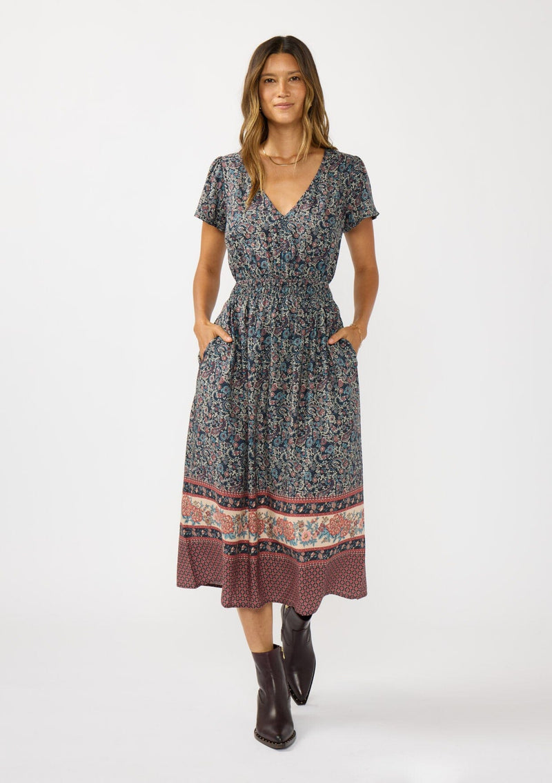 [Color: Navy/Rose] A front facing image of a brunette model wearing a navy floral border print midi dress with a classic v-neckline, short sleeves, a smocked waist, and side pockets. A fall bohemian dress perfect for any occasion. 