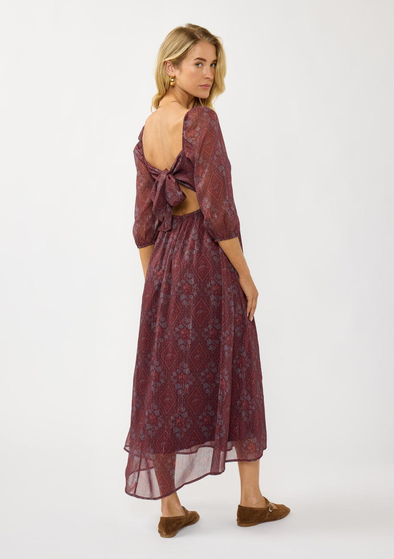 [Color: Wine/Rose] A back facing image of a blonde model wearing a floral diamond print chiffon maxi dress with metallic threads, a tie back, 3/4 sleeves, and a scoop neckline. A flattering special occasion dress for weddings or holidays. 