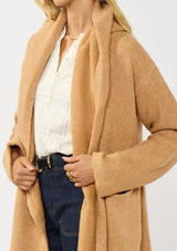 [Color: Camel] A detail image of a blonde model wearing a best selling oversized cardigan in a camel brown color. A cozy and thick sweater coat with a hood, an open front, side pockets, a cocoon style silhouette, and a mid length hem. 