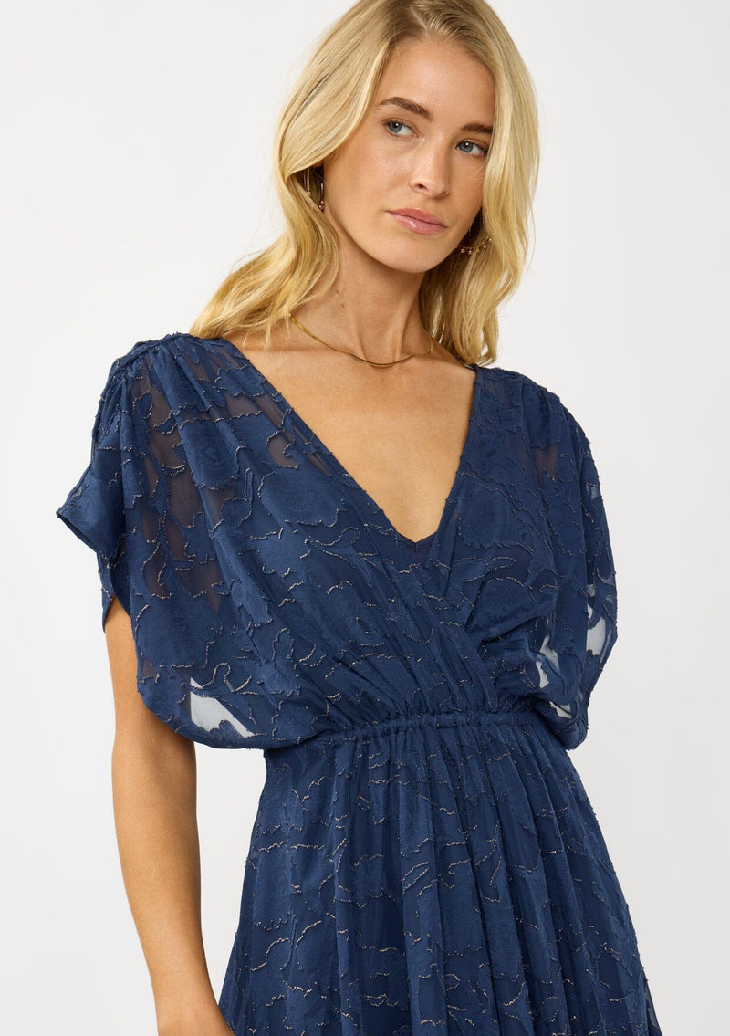 [Color: Navy] A detail image of a blonde model wearing a blue floral jacquard maxi dress with gold foil accents. Featuring a flattering surplice v-neckline, dolman sleeves, elastic waist, tie back, and a handkerchief hemline. 