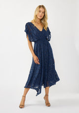 [Color: Navy] A front facing image of a blonde model wearing a blue floral jacquard maxi dress with gold foil accents. Featuring a flattering surplice v-neckline, dolman sleeves, elastic waist, tie back, and a handkerchief hemline. 
