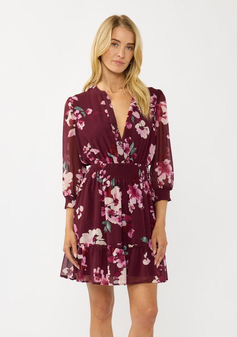 [Color: Wine/Dusty Rose] A front facing image of a blonde model wearing a burgundy floral mini dress with 3/4 sleeves, a smocked waist, button front, and a ruffle tiered skirt. 