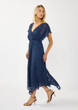 [Color: Navy] A side  facing image of a blonde model wearing a blue floral jacquard maxi dress with gold foil accents. Featuring a flattering surplice v-neckline, dolman sleeves, elastic waist, tie back, and a handkerchief hemline. 