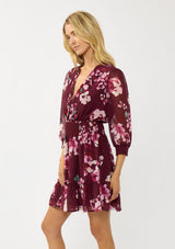 [Color: Wine/Dusty Rose] A side facing image of a blonde model wearing a burgundy floral mini dress with 3/4 sleeves, a smocked waist, button front, and a ruffle tiered skirt. 