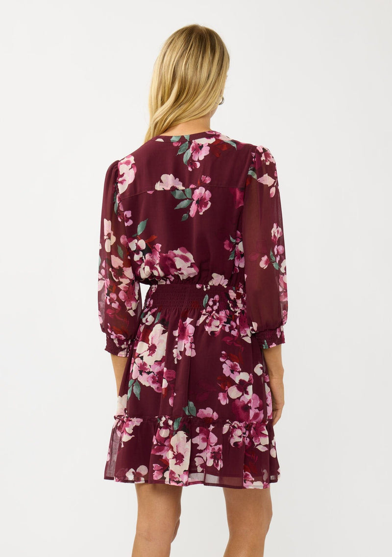 [Color: Wine/Dusty Rose] A back facing image of a blonde model wearing a burgundy floral mini dress with 3/4 sleeves, a smocked waist, button front, and a ruffle tiered skirt. 