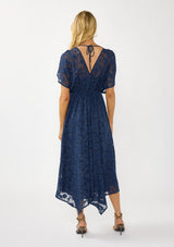 [Color: Navy] A back facing image of a blonde model wearing a blue floral jacquard maxi dress with gold foil accents. Featuring a flattering surplice v-neckline, dolman sleeves, elastic waist, tie back, and a handkerchief hemline. 