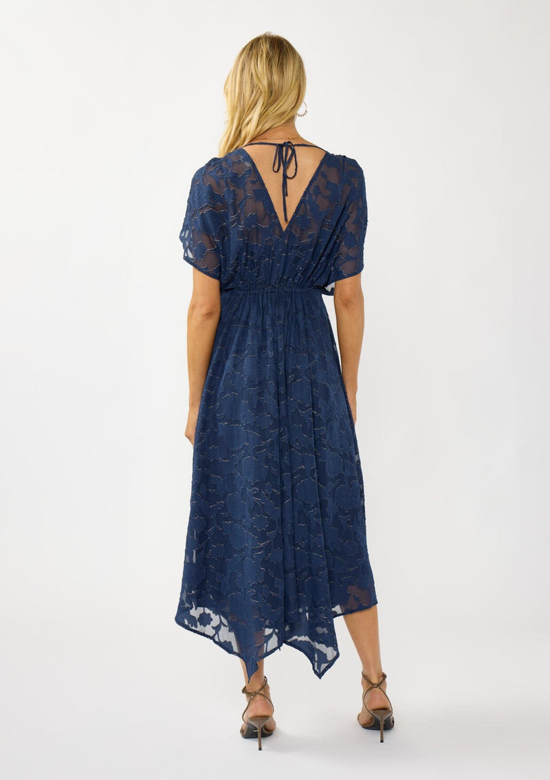 [Color: Navy] A back facing image of a blonde model wearing a blue floral jacquard maxi dress with gold foil accents. Featuring a flattering surplice v-neckline, dolman sleeves, elastic waist, tie back, and a handkerchief hemline. 