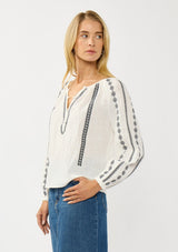 [Color: White/Black] A side facing image of a blonde model wearing a white cotton top with black embroidered details. A classic boho top with a split v-neckline, button front, and long sleeves.  