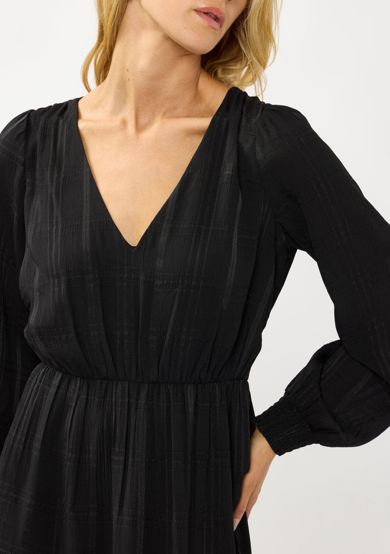 [Color: Black] A detail image of  a blonde model wearing a black plaid chiffon mini dress with a subtle sheen. Featuring a v-neckline, elastic waist, long sleeves, and a ruffle trimmed hem. A classic black mini with plaid pattern for the holidays. 
