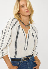 [Color: White/Black] A front facing image of a blonde model wearing a white cotton top with black embroidered details. A classic boho top with a split v-neckline, button front, and long sleeves.  