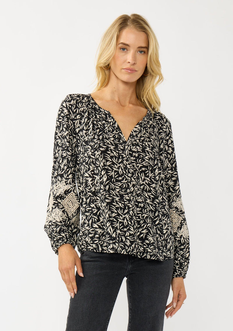 [Color: Black/Natural] A front facing image of a blonde model wearing an abstract print blouse with embroidered details, long sleeves, a button front, and a split v-neckline. 