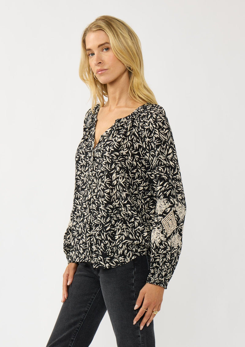 [Color: Black/Natural] A side facing image of a blonde model wearing an abstract print blouse with embroidered details, long sleeves, a button front, and a split v-neckline. 