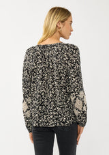 [Color: Black/Natural] A back facing image of a blonde model wearing an abstract print blouse with embroidered details, long sleeves, a button front, and a split v-neckline. 