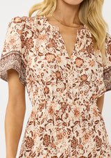 [Color: Natural/Brown] A detail image of a blonde model wearing a bohemian floral border print mini dress with a split v-neckline, short sleeves, and an elastic waist. 