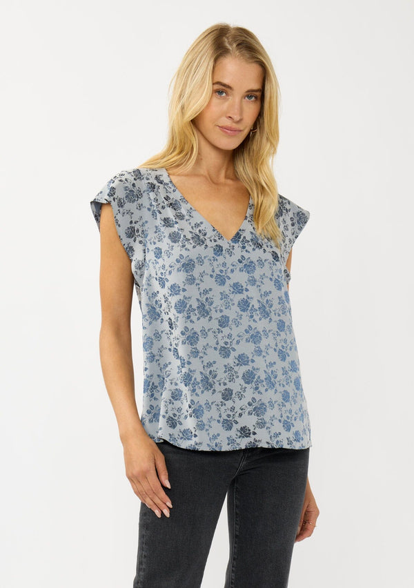 [Color: Blue] A front facing image of a blonde model wearing a blue floral top with a subtle sheen. Featuring short flutter sleeves, pleated details, and a v-neckline. A versatile fall top for casual and special occasions. 