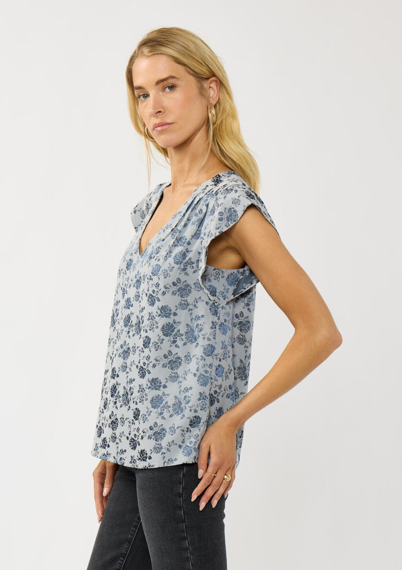 [Color: Blue] A side facing image of a blonde model wearing a blue floral top with a subtle sheen. Featuring short flutter sleeves, pleated details, and a v-neckline. A versatile fall top for casual and special occasions. 