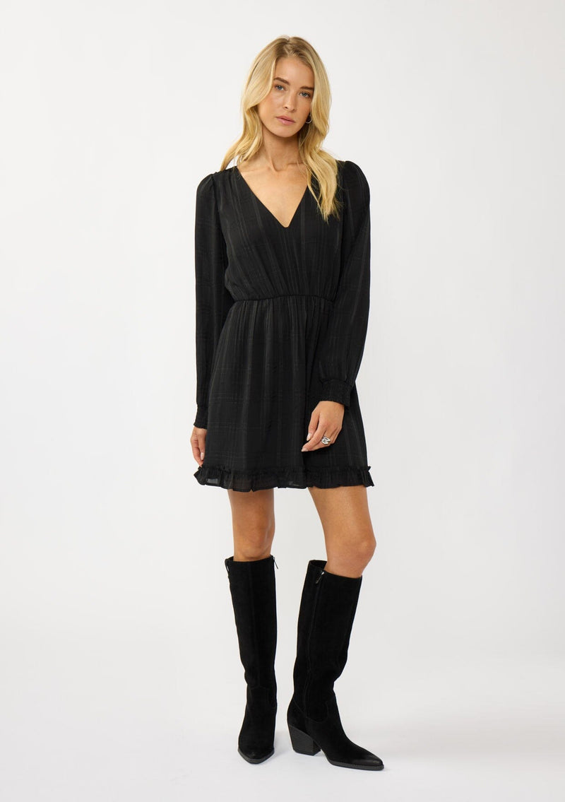 [Color: Black] A front facing image of  a blonde model wearing a black plaid chiffon mini dress with a subtle sheen. Featuring a v-neckline, elastic waist, long sleeves, and a ruffle trimmed hem. A classic black mini with plaid pattern for the holidays. 