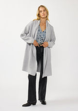 [Color: Platinum] A front facing image of a blonde model wearing a best selling oversized cardigan in a grey blue color. A cozy and thick sweater coat with a hood, an open front, side pockets, a cocoon style silhouette, and a mid length hem. 