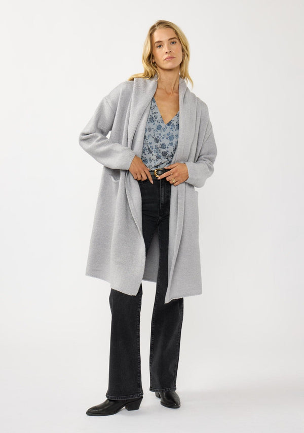 [Color: Platinum] A front facing image of a blonde model wearing a best selling oversized cardigan in a grey blue color. A cozy and thick sweater coat with a hood, an open front, side pockets, a cocoon style silhouette, and a mid length hem. 