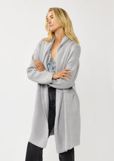 [Color: Platinum] A front facing image of a blonde model wearing a best selling oversized cardigan in a grey blue color. A cozy and thick sweater coat with a hood, an open front, side pockets, a cocoon style silhouette, and a mid length hem. 