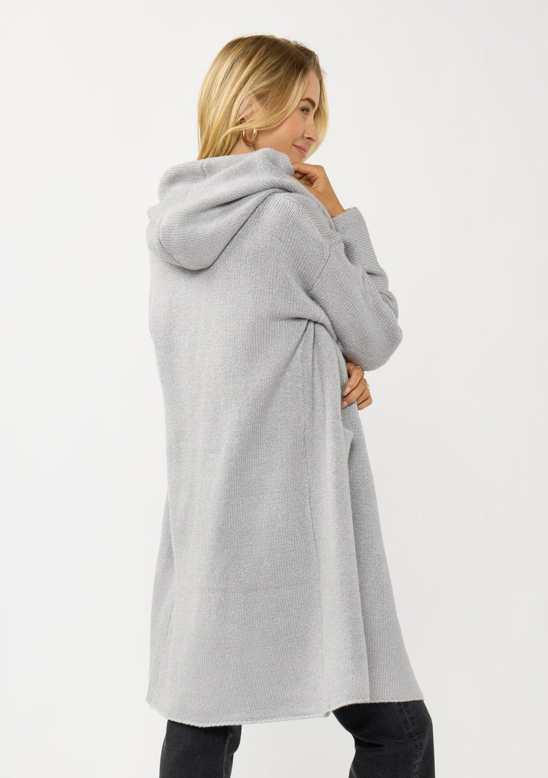 [Color: Platinum] A back facing image of a blonde model wearing a best selling oversized cardigan in a grey blue color. A cozy and thick sweater coat with a hood, an open front, side pockets, a cocoon style silhouette, and a mid length hem. 
