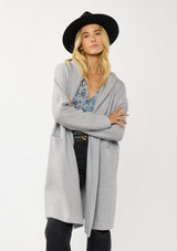 [Color: Platinum] A front facing image of a blonde model wearing a best selling oversized cardigan in a grey blue color. A cozy and thick sweater coat with a hood, an open front, side pockets, a cocoon style silhouette, and a mid length hem. 