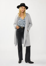 [Color: Platinum] A front facing image of a blonde model wearing a best selling oversized cardigan in a grey blue color. A cozy and thick sweater coat with a hood, an open front, side pockets, a cocoon style silhouette, and a mid length hem. 