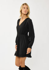 [Color: Black] A side facing image of  a blonde model wearing a black plaid chiffon mini dress with a subtle sheen. Featuring a v-neckline, elastic waist, long sleeves, and a ruffle trimmed hem. A classic black mini with plaid pattern for the holidays. 