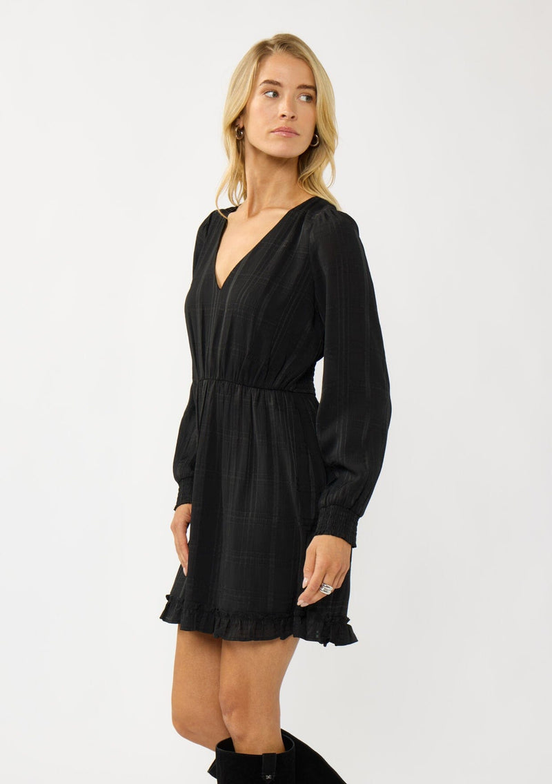 [Color: Black] A side facing image of  a blonde model wearing a black plaid chiffon mini dress with a subtle sheen. Featuring a v-neckline, elastic waist, long sleeves, and a ruffle trimmed hem. A classic black mini with plaid pattern for the holidays. 