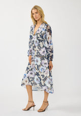 [Color: Grey/Navy] A side facing image of a blonde model wearing a grey and purple floral midi dress with a high-low hemline, empire waist, long sleeves, and a deep v-neckline. Feminine and elegant for weddings and special occasions. 