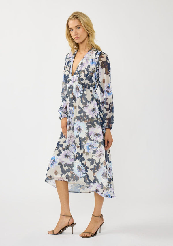 [Color: Grey/Navy] A side facing image of a blonde model wearing a grey and purple floral midi dress with a high-low hemline, empire waist, long sleeves, and a deep v-neckline. Feminine and elegant for weddings and special occasions. 