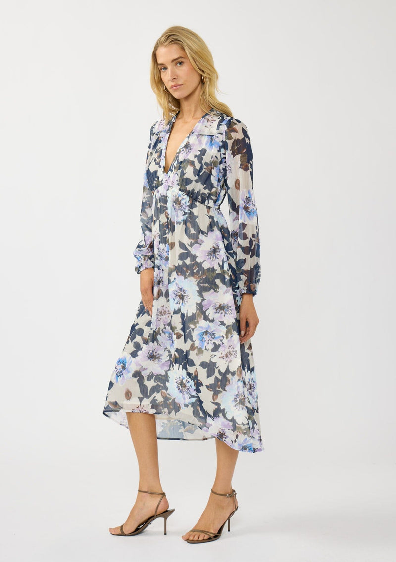 [Color: Grey/Navy] A side facing image of a blonde model wearing a grey and purple floral midi dress with a high-low hemline, empire waist, long sleeves, and a deep v-neckline. Feminine and elegant for weddings and special occasions. 