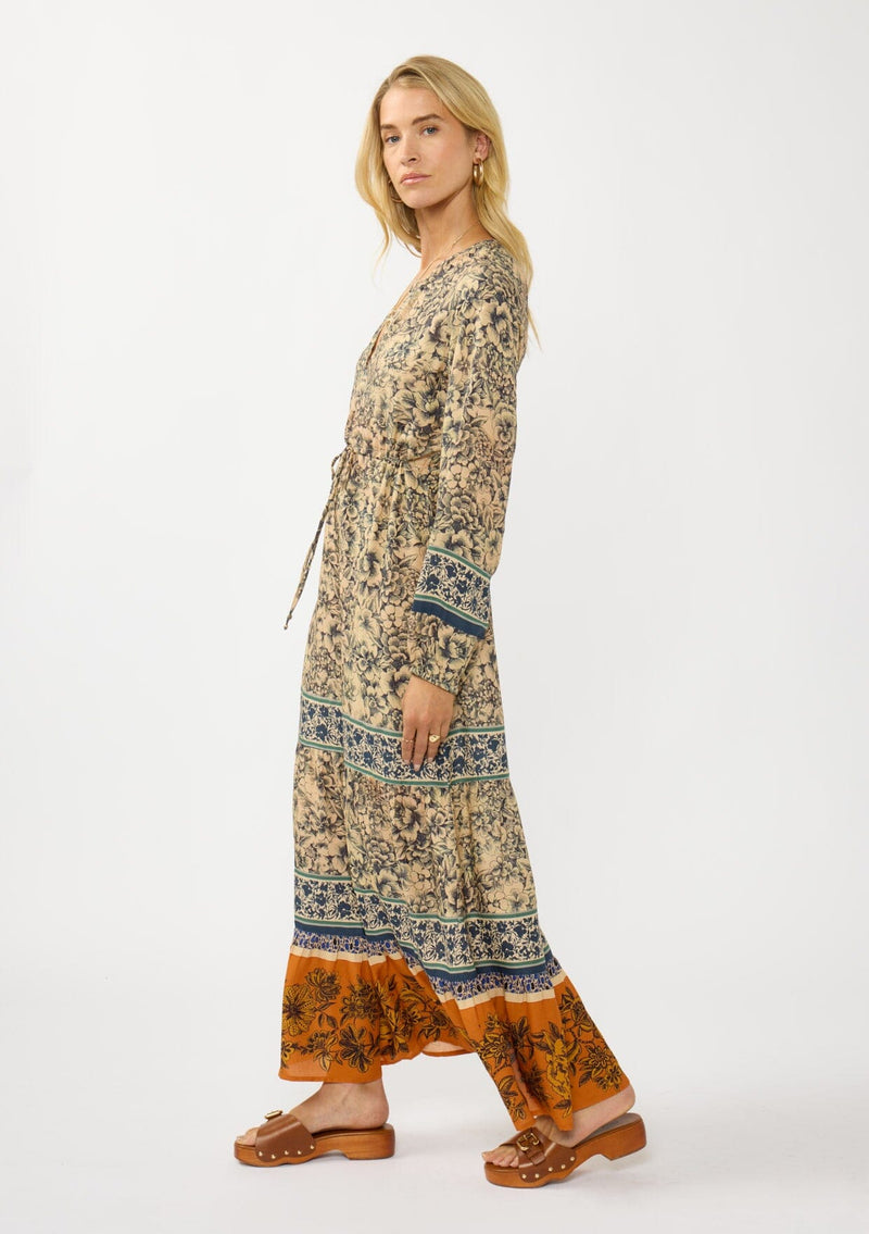 [Color: Natural/Rust] A side facing image of a blonde model wearing a yellow and orange floral border print maxi dress. Featuring long sleeves, a v-neckline, drawstring waist, and a tiered skirt. A fall boho dress for any occasion. 