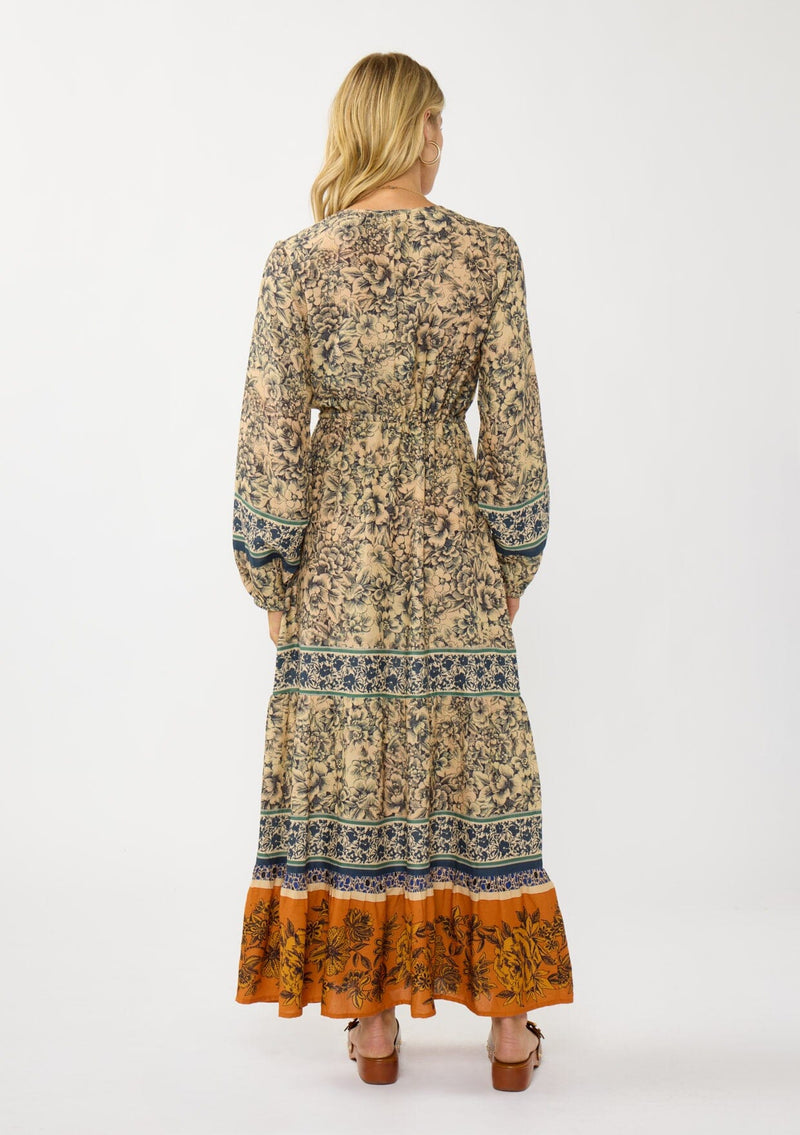 [Color: Natural/Rust] A back facing image of a blonde model wearing a yellow and orange floral border print maxi dress. Featuring long sleeves, a v-neckline, drawstring waist, and a tiered skirt. A fall boho dress for any occasion. 