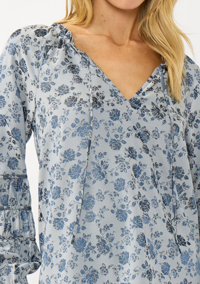 [Color: Blue] A detail image of a blonde model wearing a blue floral blouse with a subtle sheen. Featuring long sleeves with shirred details, a split v-neckline with tie detail, and a ruffled neckline. 