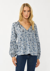 [Color: Blue] A front facing image of a blonde model wearing a blue floral blouse with a subtle sheen. Featuring long sleeves with shirred details, a split v-neckline with tie detail, and a ruffled neckline. 