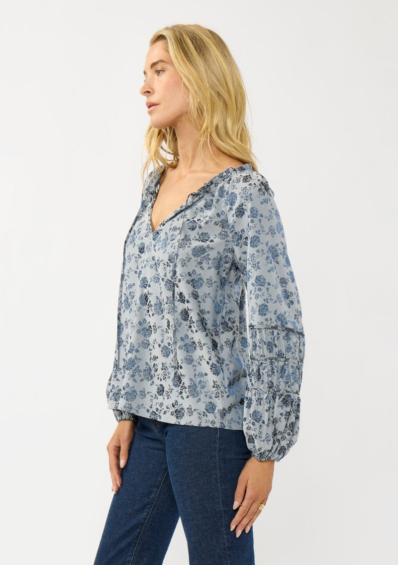 [Color: Blue] A side facing image of a blonde model wearing a blue floral blouse with a subtle sheen. Featuring long sleeves with shirred details, a split v-neckline with tie detail, and a ruffled neckline. 