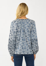 [Color: Blue] A back facing image of a blonde model wearing a blue floral blouse with a subtle sheen. Featuring long sleeves with shirred details, a split v-neckline with tie detail, and a ruffled neckline. 