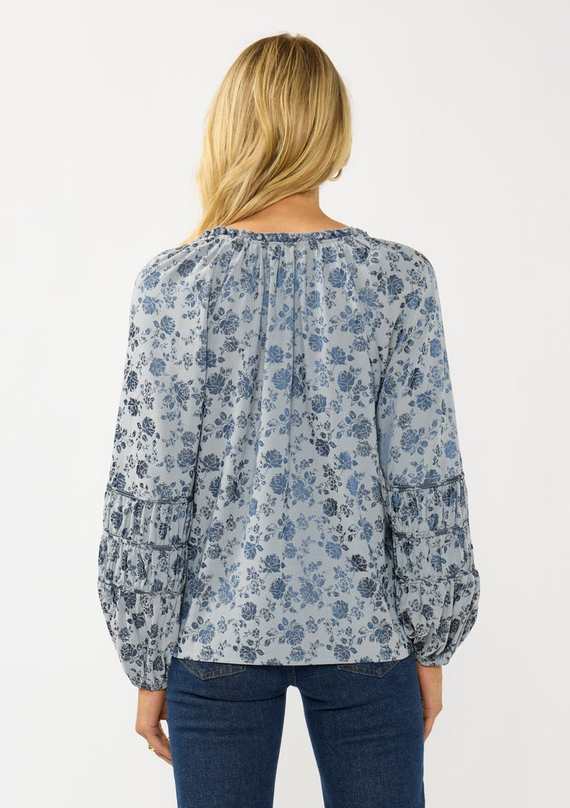 [Color: Blue] A back facing image of a blonde model wearing a blue floral blouse with a subtle sheen. Featuring long sleeves with shirred details, a split v-neckline with tie detail, and a ruffled neckline. 