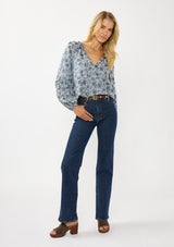 [Color: Blue] A front facing image of a blonde model wearing a blue floral blouse with a subtle sheen. Featuring long sleeves with shirred details, a split v-neckline with tie detail, and a ruffled neckline. 