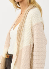 [Color: Natural/Tan] A detail image of a blonde model wearing a pink and beige cable knit patchwork cardigan with an open front and cozy long sleeves. A cozy layering piece for the fall season. 