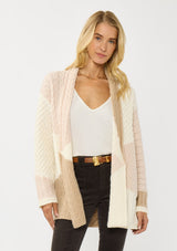 [Color: Natural/Tan] A front facing image of a blonde model wearing a pink and beige cable knit patchwork cardigan with an open front and cozy long sleeves. A cozy layering piece for the fall season. 