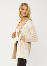 [Color: Natural/Tan] A side facing image of a blonde model wearing a pink and beige cable knit patchwork cardigan with an open front and cozy long sleeves. A cozy layering piece for the fall season. 